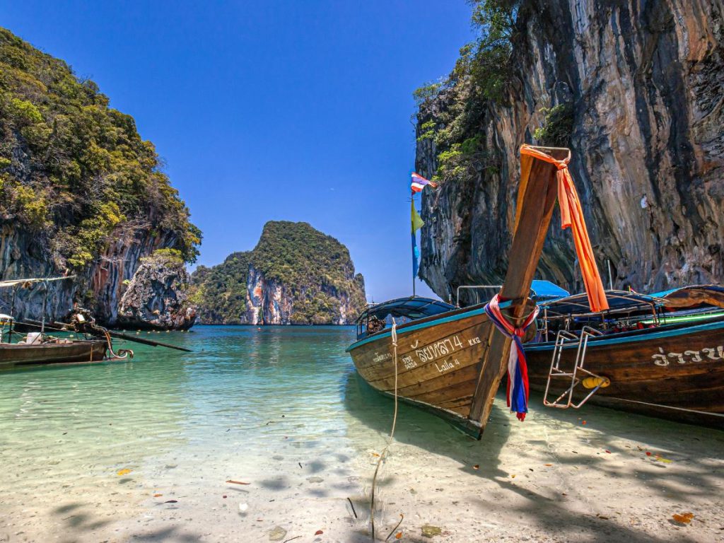 Tipiche imbarcazioni "long-tail" tailandesi ormeggiate su una spiaggia di sabbia bianca con acque cristalline, circondate da alte scogliere calcaree ricoperte di vegetazione