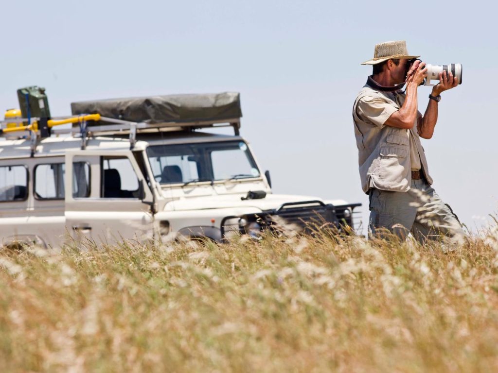 Un uomo con un cappello di paglia e un gilet multitasche punta un obiettivo fotografico professionale mentre si trova accanto a un fuoristrada bianco in un campo aperto.
