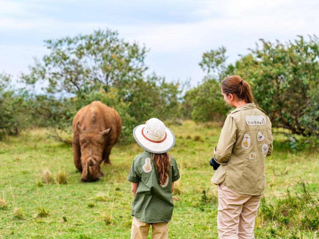 Una donna e una bambina di spalle, vestite con abbigliamento tecnico da safari decorato con patch di animali, osservano un rinoceronte in un prato verde. Utile per sapere come vestirsi per un safari.