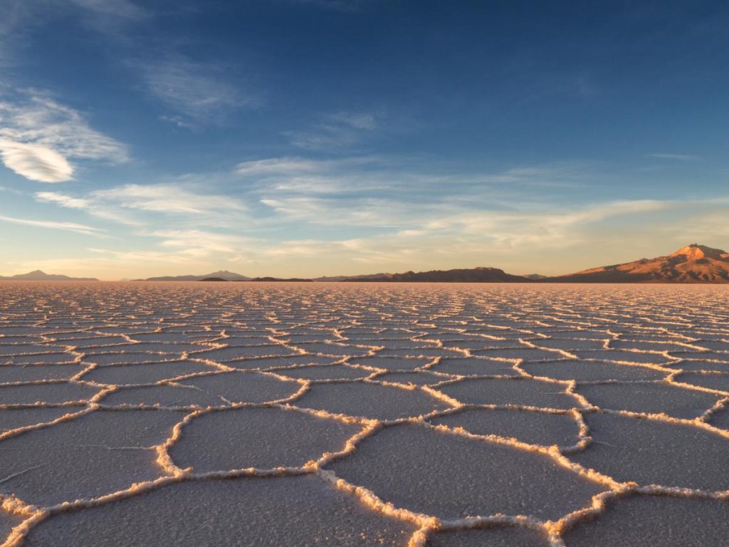 Veduta prospettica del Salar de Uyuni al tramonto, che mostra la caratteristica superficie di sale bianco screpolata in formazioni esagonali che si estendono verso le montagne all'orizzonte sotto un cielo blu sfumato.