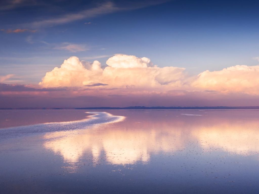 Paesaggio surreale del Salar de Uyuni coperto da un sottile strato d'acqua, che crea un perfetto effetto specchio. Grandi nuvole bianche e rosate si riflettono sulla superficie immobile durante l'ora d'oro.