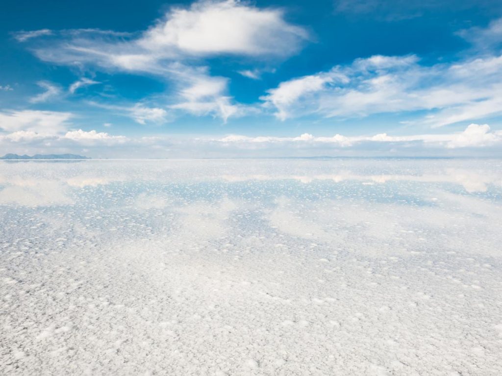 Una distesa luminosa e infinita di sale bianco ricoperta d'acqua, dove il cielo azzurro e le nuvole bianche si fondono perfettamente con il terreno, eliminando la linea dell'orizzonte.