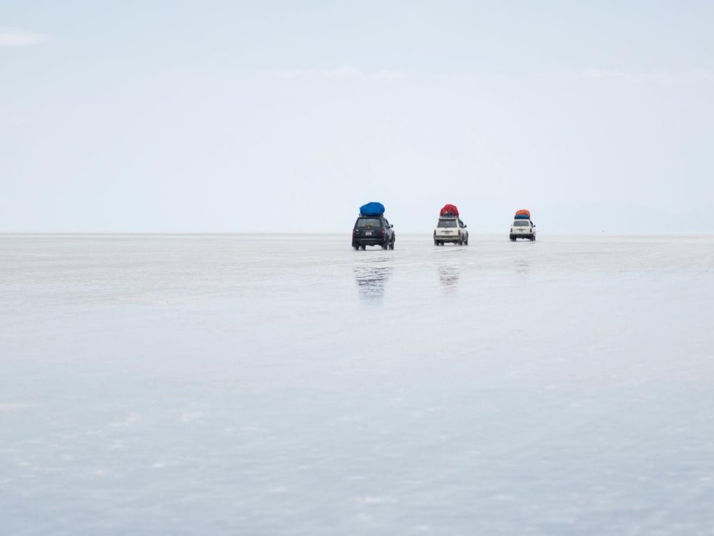 Tre fuoristrada con bagagli colorati sul tetto che guidano in fila attraverso l'immensa e piatta distesa bianca del Salar de Uyuni, apparendo come piccoli punti in un paesaggio bianco e minimalista.