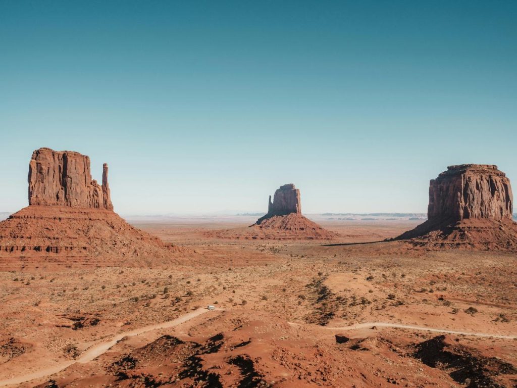 Una vasta panoramica della Monument Valley sotto un cielo azzurro limpido, durante un viaggio di turismo d'avventura negli Stati Uniti. In primo piano si snoda una strada sterrata che attraversa il terreno desertico rossastro, punteggiato da bassi arbusti, con le iconiche formazioni rocciose a forma di "butte" che si stagliano imponenti all'orizzonte.