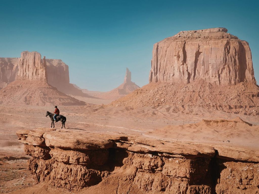 Un uomo a cavallo, con cappello a tesa larga e camicia rossa, osserva il panorama della Monument Valley dal bordo di un precipizio roccioso. Sullo sfondo, le maestose formazioni di arenaria emergono da una leggera foschia dorata sotto il sole.
