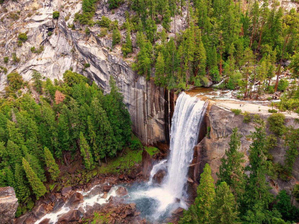 Una veduta aerea di una potente cascata che si tuffa da un'alta parete granitica nel Parco Nazionale di Yosemite. La cascata è circondata da una fitta foresta di pini verdi e rocce scoscese, con l'acqua che si infrange in una nuvola di spruzzi bianchi alla base.