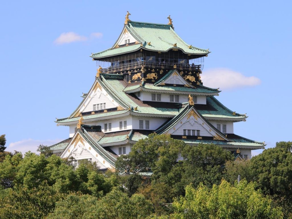 Veduta dal basso del Castello di Osaka in Giappone, circondato da cime di alberi verdi e rigogliosi sotto un cielo azzurro con poche nuvole. L'edificio storico mostra la tipica architettura giapponese a più livelli con tetti verde acqua dalle punte rivolte verso l'alto, pareti bianche e dettagli decorativi dorati e neri sulla facciata superiore. Da vedere durante un viaggio su misura tra Giappone e Fiji.
