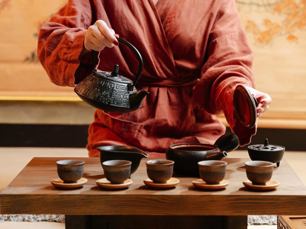 Primo piano ravvicinato delle mani di una persona che indossa una veste color ruggine durante una cerimonia del tè tradizionale. La mano sinistra solleva una teiera in ghisa nera decorata mentre versa il tè in una serie di cinque piccole tazze di ceramica scura disposte in fila su un tavolo di legno scuro.