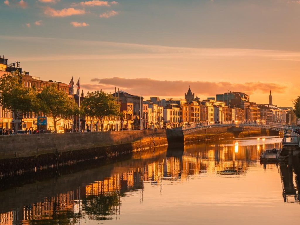 Scorcio di Dublino al tramonto con il fiume Liffey che riflette la luce dorata del sole, i palazzi storici della città e l'Ha'penny Bridge sullo sfondo.