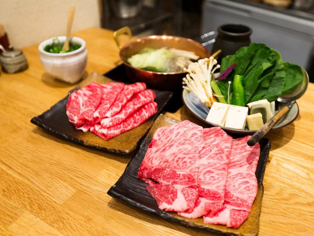 Un set da tavola per shabu-shabu o sukiyaki con due piatti di carne di manzo Wagyu finemente marmorizzata, una ciotola di verdure fresche (funghi enoki, tofu e foglie verdi) e una pentola di rame fumante su un tavolo di legno.
