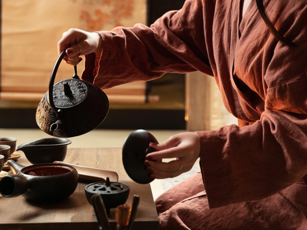Un dettaglio ravvicinato di una persona che indossa un kimono marrone mentre versa l'acqua da una teiera in ghisa nera finemente decorata. Sul tavolo di legno sono disposti diversi utensili tradizionali per la cerimonia del tè, tra cui piccole tazze e contenitori in ceramica.