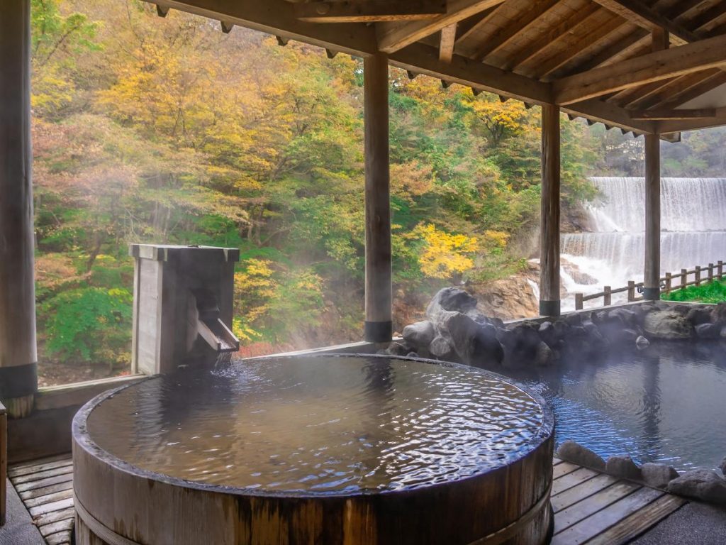 Una vasca termale all'aperto (rotenburo) realizzata in legno circolare, colma di acqua calda fumante. La vasca è situata sotto un porticato in legno che si affaccia su un bosco con colori autunnali e una cascata a più livelli sullo sfondo.
