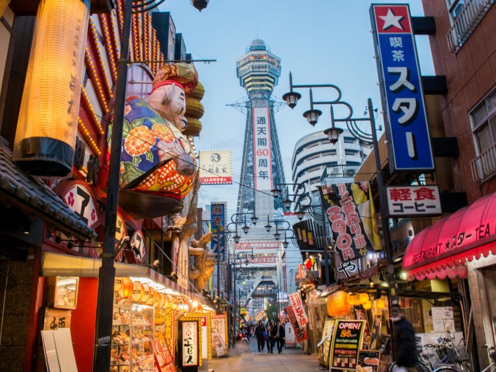 Una vivace strada pedonale nel quartiere Shinsekai di Osaka al crepuscolo, affollata di insegne colorate e lanterne, con l'iconica torre Tsutenkaku che svetta sullo sfondo.