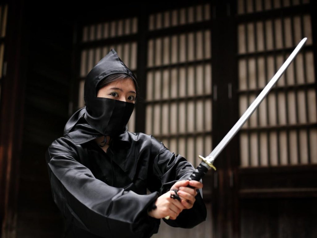 Una persona vestita con una divisa nera da ninja e il volto parzialmente coperto, che impugna una katana con entrambe le mani in una posa di guardia davanti a una tradizionale porta scorrevole giapponese.