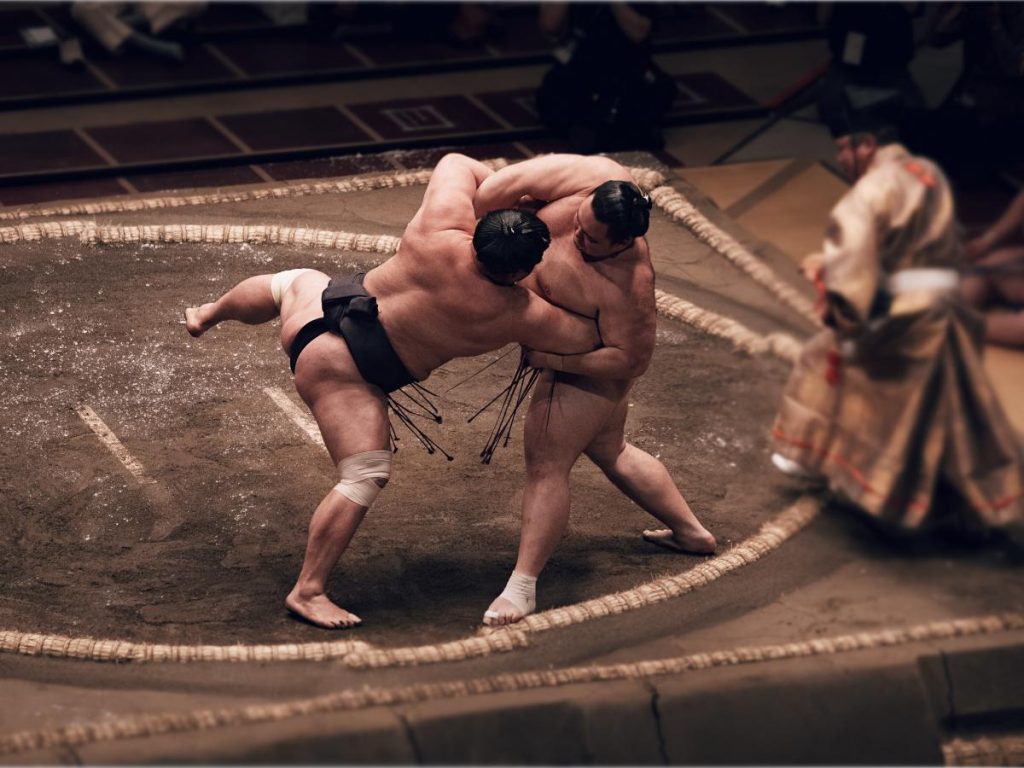 Due lottatori di sumo impegnati in un combattimento all'interno del dohyo (ring di sabbia), circondati dal bordo di paglia, con un arbitro (gyoji) in abiti tradizionali visibile lateralmente.