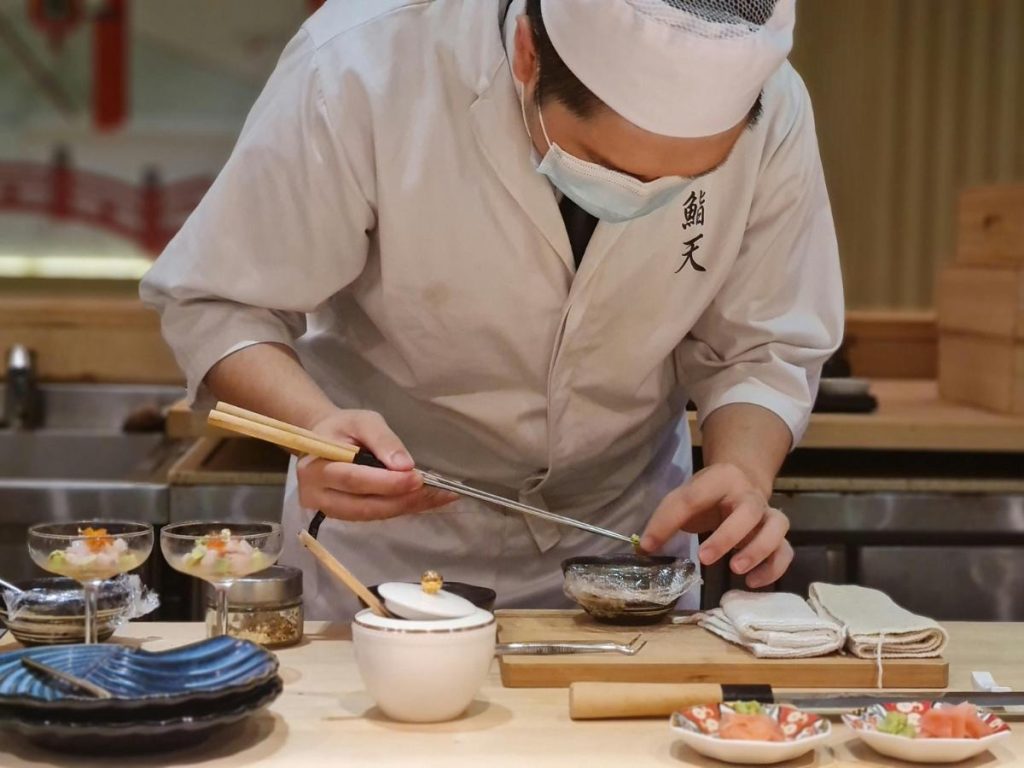 Uno chef di sushi in divisa bianca e mascherina mentre prepara con precisione millimetrica dei piatti gourmet utilizzando delle lunghe pinze da cucina. Davanti a lui, sul bancone in legno, sono disposti piccoli piattini con ingredienti freschi, salse e guarnizioni.