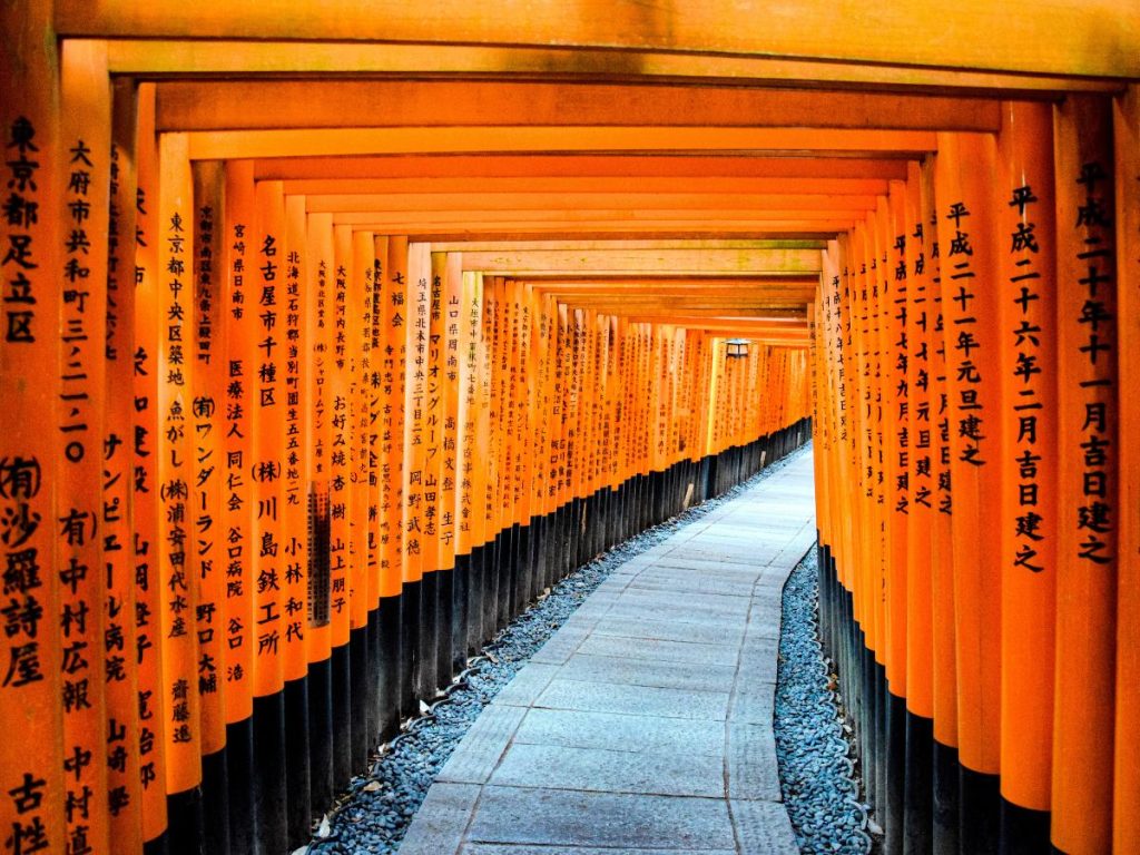 Prospettiva interna del tunnel di pilastri torii arancioni e neri del santuario di Fushimi Inari-taisha a Kyoto. I pali di legno sono disposti fittamente uno dopo l'altro lungo un sentiero lastricato curvo, e presentano iscrizioni con caratteri giapponesi neri incisi sulla loro superficie.
