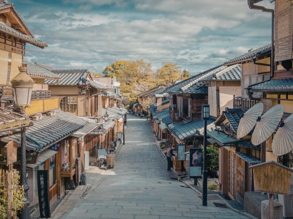Veduta dall'alto di una pittoresca e tranquilla strada acciottolata in discesa nel quartiere storico di Gion a Kyoto. La via è fiancheggiata da tradizionali case giapponesi a due piani in legno con tetti in tegole scure, lanterne d'epoca e ombrelli di carta aperti esposti all'esterno di un negozio. Da vedere durante un viaggio su misura tra Giappone e Fiji.