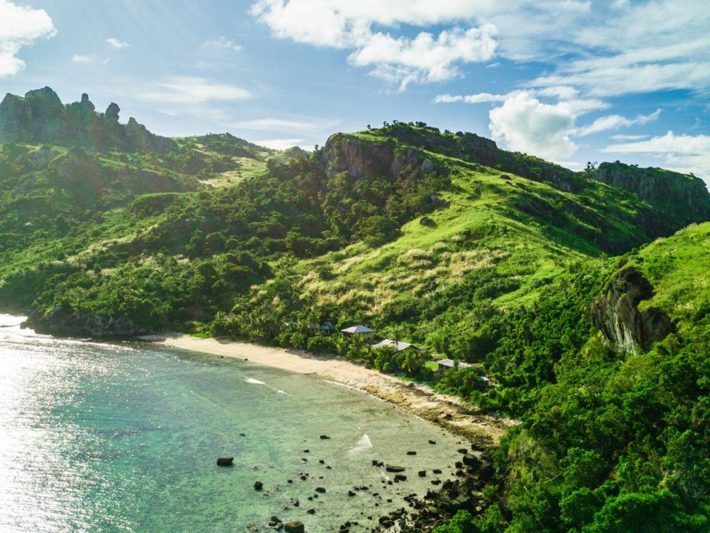 Veduta aerea panoramica di una baia tropicale alle isole Fiji. Una spiaggia di sabbia chiara a forma di mezzaluna è bagnata da acque verdi e trasparenti, mentre l'entroterra è dominato da colline vulcaniche ricoperte da una fitta e lussureggiante vegetazione verde sotto un cielo parzialmente nuvoloso.