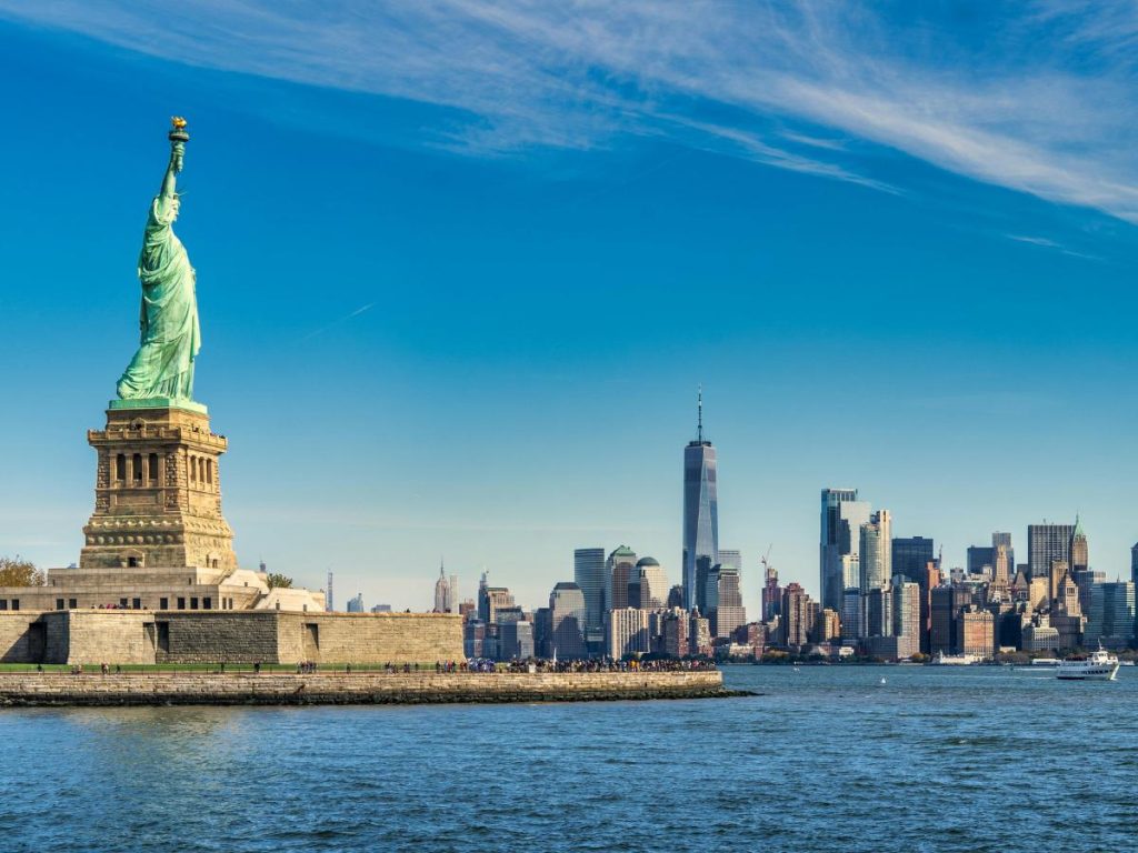 La Statua della Libertà in primo piano a sinistra con lo skyline di Manhattan, il One World Trade Center e il fiume Hudson sullo sfondo a New York, da vedere durante un viaggio in nord America con scalo a Dublino.