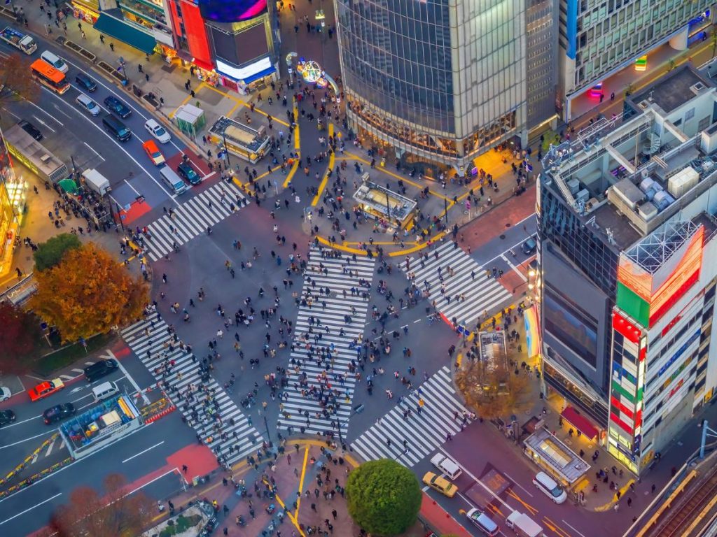 Veduta aerea zenitale del celebre incrocio pedonale di Shibuya a Tokyo. Numerose strisce pedonali bianche attraversano l'asfalto grigio in varie direzioni, popolate da centinaia di pedoni in movimento. Ai lati si vedono i tetti degli edifici urbani, maxischermi pubblicitari e strade con automobili e autobus.