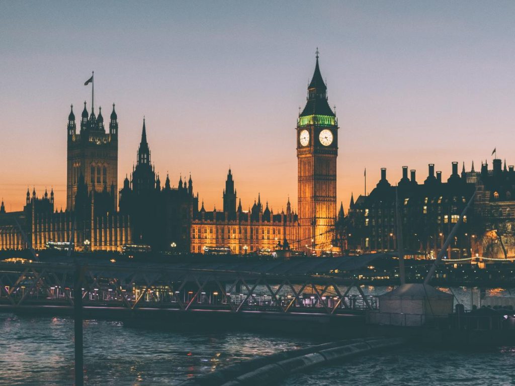 Il Palazzo di Westminster e l'iconica torre dell'orologio del Big Ben a Londra illuminati al crepuscolo, visti dall'altra parte del fiume Tamigi. L'orizzonte mostra le sagome degli edifici storici stagliate contro un cielo sfumato dall'arancione al blu scuro. In primo piano, le acque del fiume riflettono le luci della città e si nota la struttura di un ponte pedonale.
