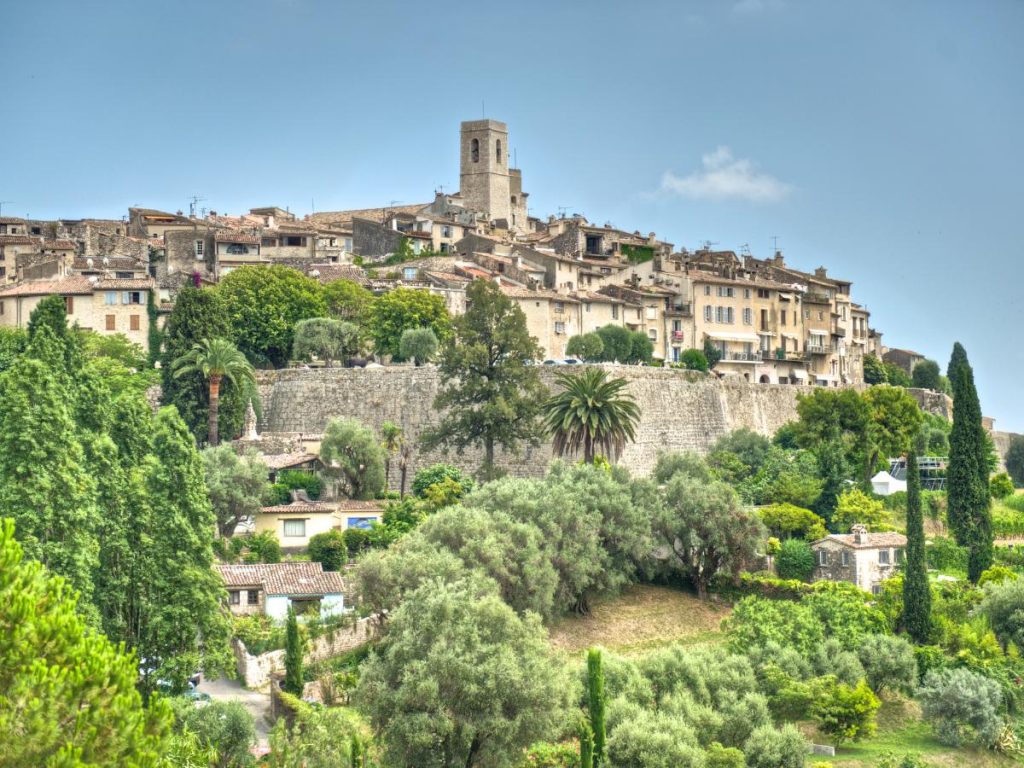 Veduta di un antico borgo medievale arroccato su una collina (Saint-Paul-de-Vence). Le case in pietra dalle tonalità chiare e dai tetti marroni si sviluppano attorno a una torre campanaria centrale. La collina è circondata da imponenti mura fortificate e digrada verso il basso con una ricca vegetazione di alberi verdi e palme.