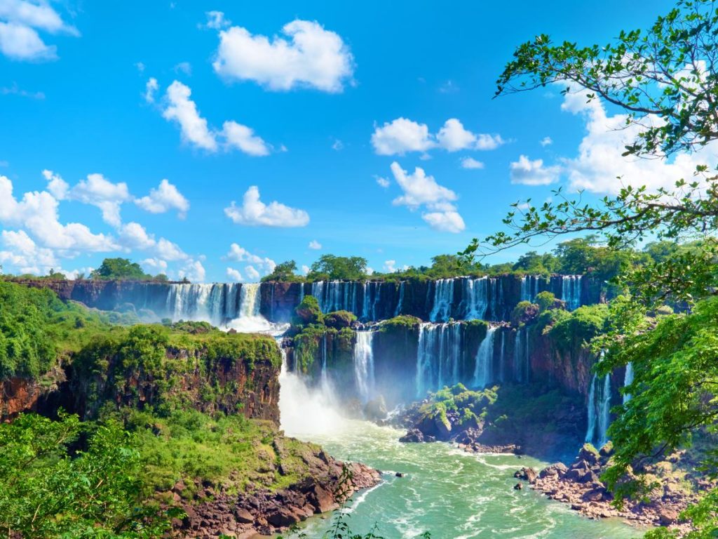 Ampia veduta panoramica delle Cascate dell'Iguazù immerse in una lussureggiante vegetazione tropicale sotto un cielo azzurro con nuvole bianche.