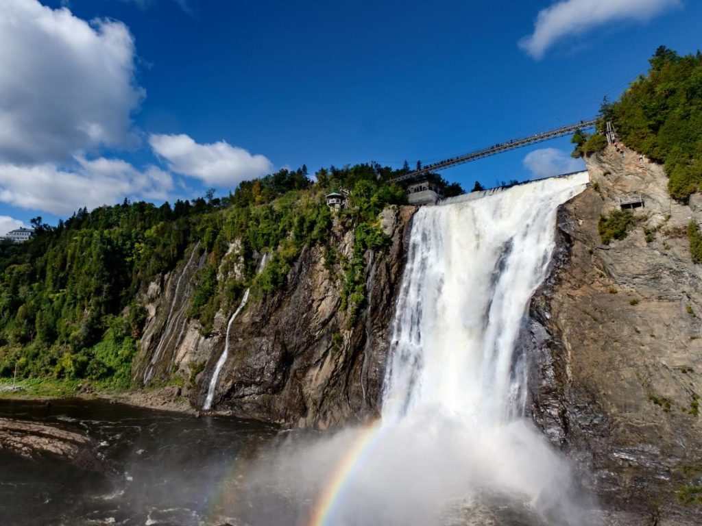 L'imponente cascata di Montmorency che si getta con forza in un bacino d'acqua, sollevando una nuvola di vapore dove si intravede un arcobaleno. Sulla cima della cascata si sviluppa un lungo ponte sospeso pedonale che collega le due sponde rocciose e boscose. Il cielo è di un azzurro intenso con nuvole bianche.