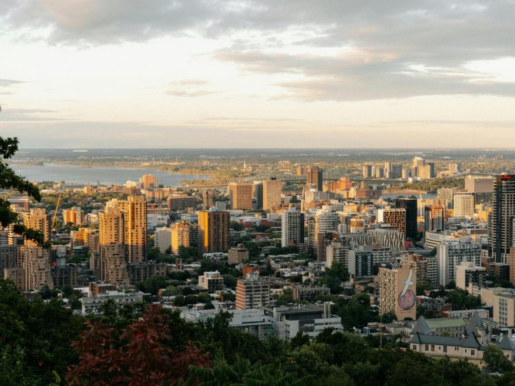 Panorama della città di Montréal vista dall'alto di una collina alberata al tramonto. La fitta distesa di grattacieli ed edifici urbani è illuminata dalla luce calda del sole calante, mentre sullo sfondo si intravedono un ampio specchio d'acqua e un ponte in lontananza sotto un cielo parzialmente nuvoloso.