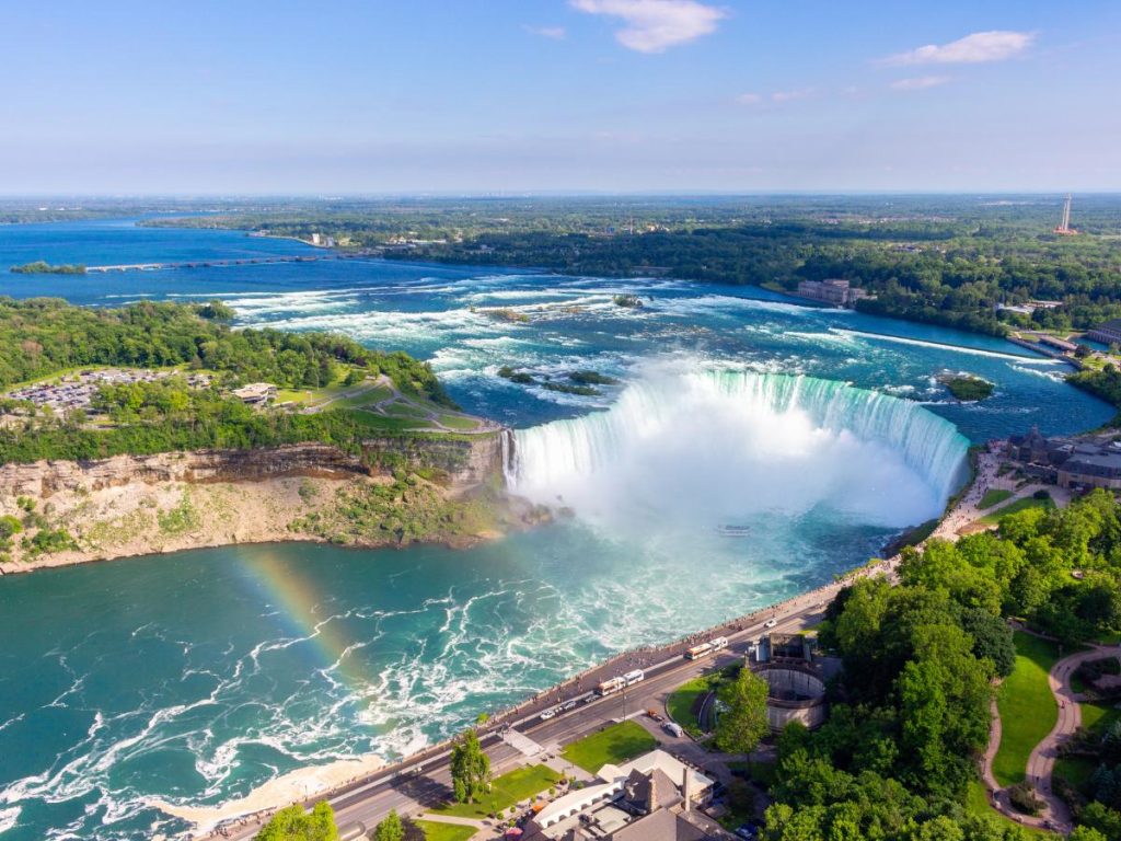 Ampia veduta aerea panoramica delle cascate del Niagara (Horseshoe Falls). L'acqua scorre rapida prima di precipitare nel profondo baratro a semicerchio, creando una grande nuvola di fumo bianco alla base. In basso a sinistra, un nitido arcobaleno attraversa le acque agitate del fiume. La riva canadese in primo piano mostra parchi verdi, una strada e strutture turistiche.