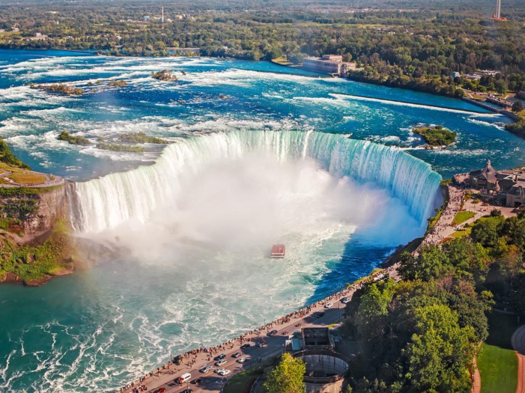 Veduta aerea ravvicinata delle cascate Horseshoe Falls a Niagara. L'imponente cascata a forma di ferro di cavallo mostra acque turchesi che si infrangono sollevando una fitta nebbia bianca, con un piccolo battello turistico che naviga vicino alla base. Sul lato destro si snoda una strada panoramica costeggiata da alberi e percorsa da pedoni e auto.