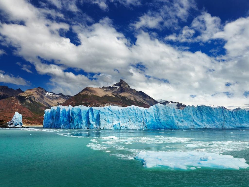 L'imponente parete di ghiaccio azzurro del ghiacciaio Perito Moreno che si staglia sulle acque turchesi del lago, con montagne e nuvole nel cielo.