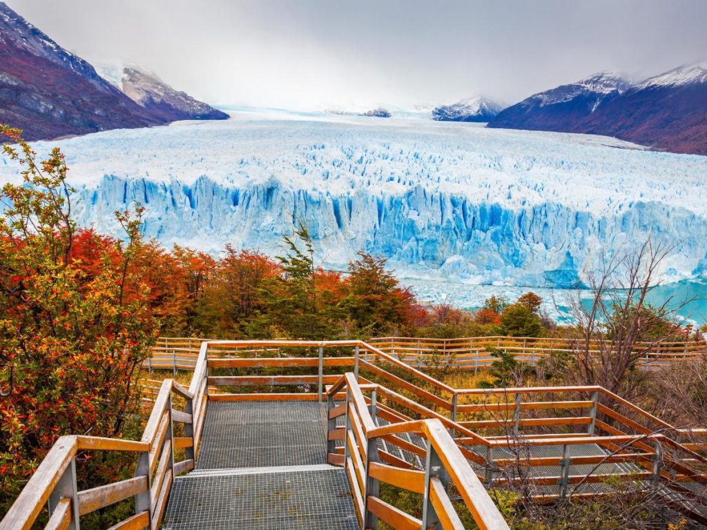 Vista panoramica del Ghiacciaio Perito Moreno da una passerella in legno, circondata da alberi con foglie autunnali rosse e arancioni sotto un cielo nuvoloso.