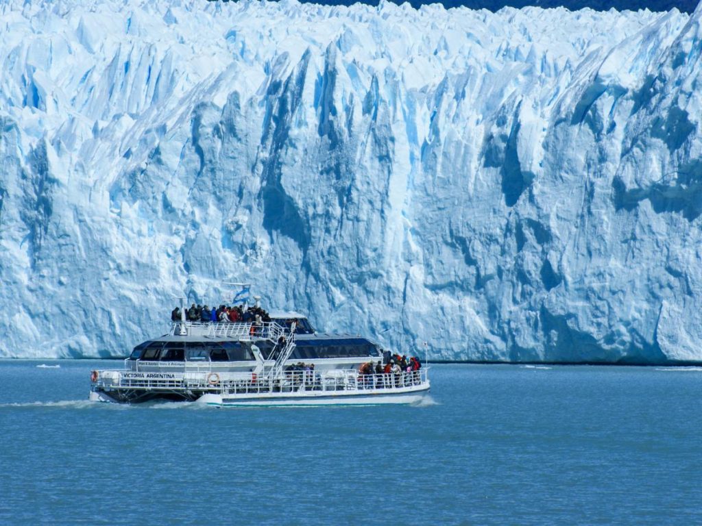 Un catamarano turistico pieno di passeggeri naviga sul lago davanti alla gigantesca e frastagliata parete di ghiaccio azzurro del ghiacciaio Perito Moreno.