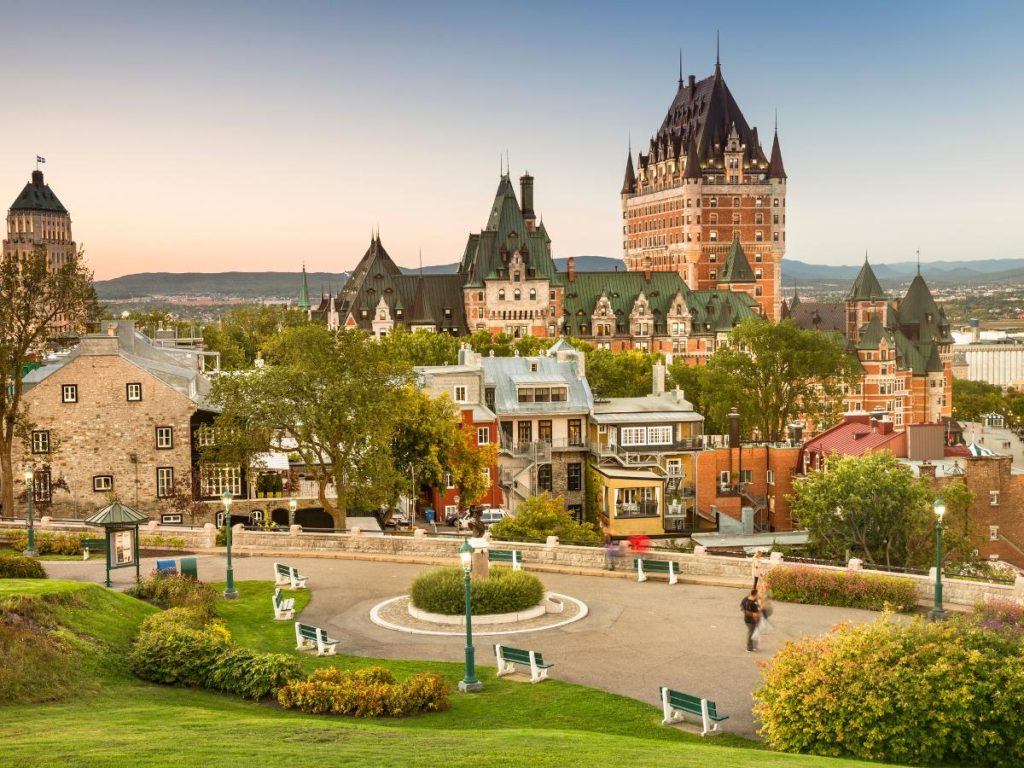 Fotografia panoramica di Québec City in Canada orientale. In primo piano si vede una terrazza pedonale con panchine verdi e lampioni circondata da prati curati. Sullo sfondo, l'imponente architettura in stile castello del Fairmont Le Château Frontenac domina la scena sotto un cielo sfumato dal tramonto.