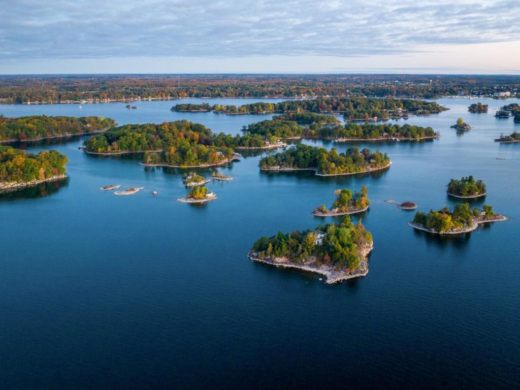Veduta aerea panoramica della regione delle Thousand Islands (Mille Isole) al crepuscolo. Numerosi isolotti ricoperti di alberi con i colori autunnali e piccole case o cottage galleggiano sulle acque calme e blu scuro del fiume. All'orizzonte si estende una linea di costa boscosa sotto un cielo leggermente nuvoloso.