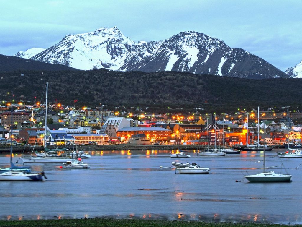 Vista panoramica serale della città di Ushuaia illuminata, con barche a vela ormeggiate nella baia e imponenti montagne innevate sullo sfondo.