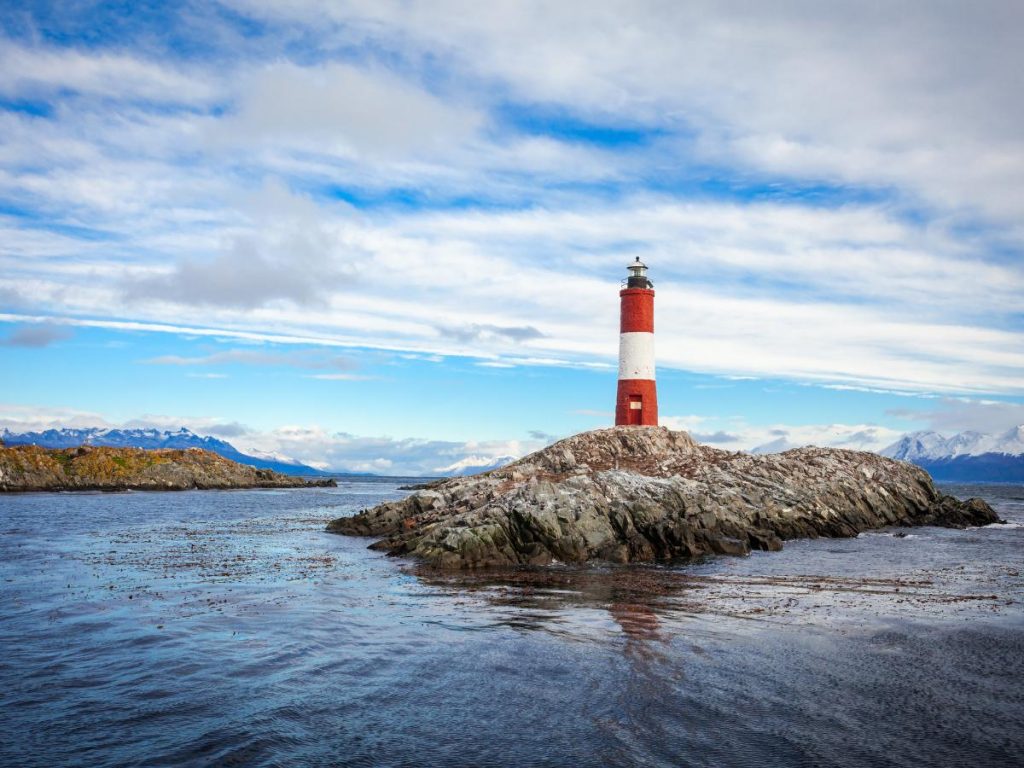 Il faro di Les Éclaireurs, a strisce rosse e bianche, situato su un isolotto roccioso nel Canale di Beagle, con montagne innevate sullo sfondo.