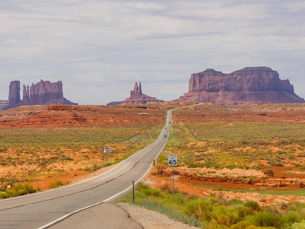 Una lunga strada asfaltata a due corsie si estende dritta e deserta verso il paesaggio desertico della Monument Valley, sotto un cielo parzialmente nuvoloso. Ai lati della strada si estende un terreno arido e rossastro con bassa vegetazione. Sulla destra è visibile un cartello stradale con il limite di velocità di 65 miglia orarie, mentre sullo sfondo si stagliano le iconiche formazioni rocciose di arenaria rossa. Da vedere durante un viaggio in America con scalo a Londra.