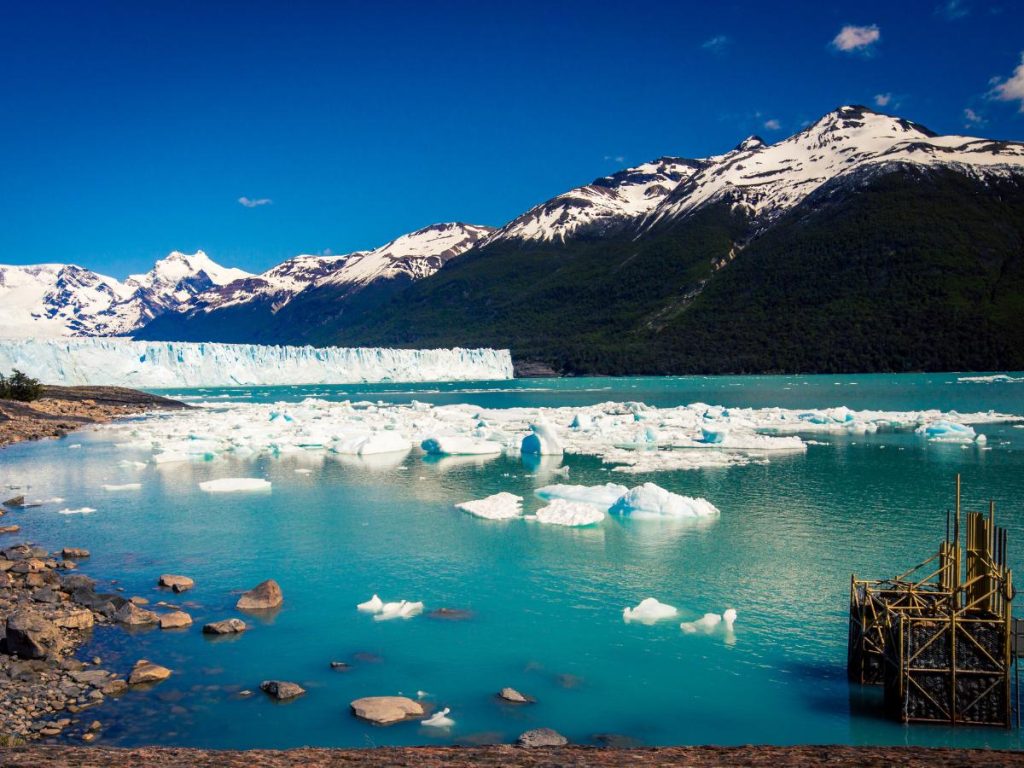 Panorama del ghiacciaio Perito Moreno in Argentina, con un muro di ghiaccio azzurro sullo sfondo, iceberg che galleggiano sulle acque turchesi del lago Argentino e montagne innevate sotto un cielo blu limpido. Da vedere durante un viaggio in Argentina.