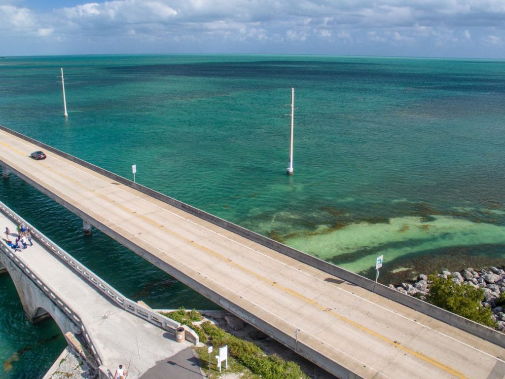 Veduta dall'alto del Seven Mile Bridge nelle Florida Keys, con un'auto in transito sul ponte stradale sopra un mare cristallino dalle tonalità verdi e blu.