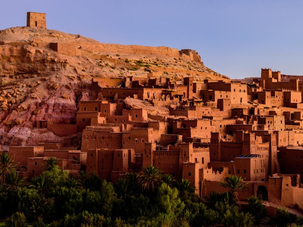Vista panoramica del ksar fortificato di Aït-Ben-Haddou illuminato dalla luce calda del tramonto con palme in primo piano.