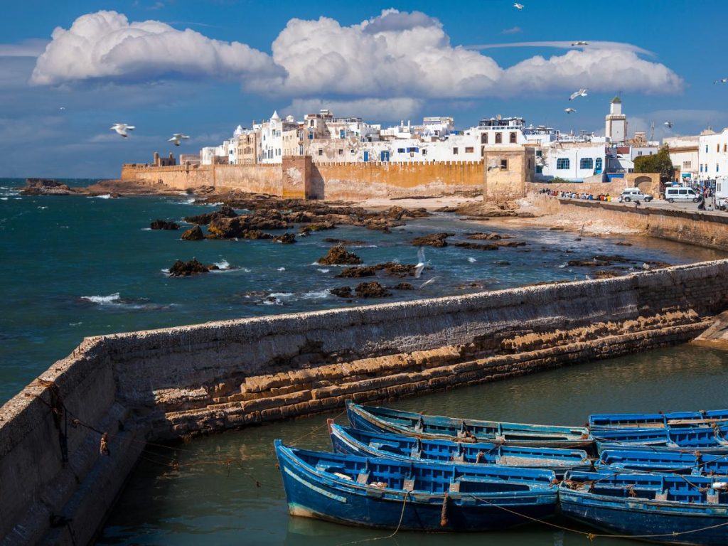 Barche da pesca blu ormeggiate nel porto di Essaouira con le mura storiche della città e il mare Atlantico sullo sfondo.