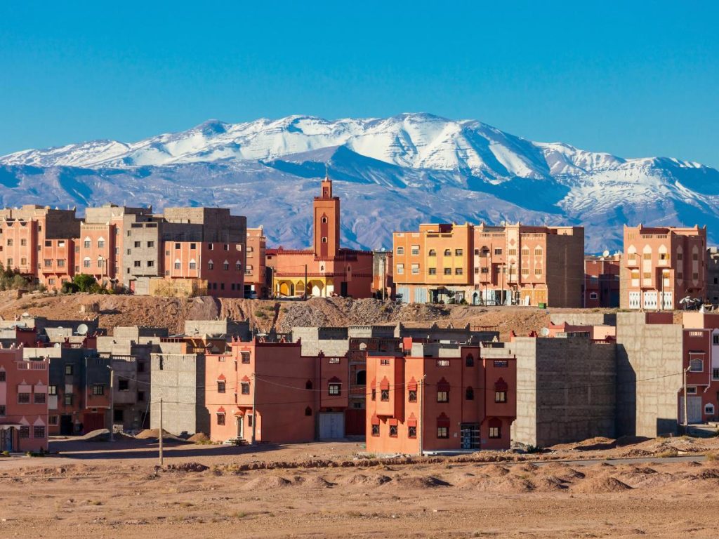Architettura tradizionale in terracotta di Ouarzazate con un minareto e l'imponente catena montuosa dell'Atlante innevata sullo sfondo.