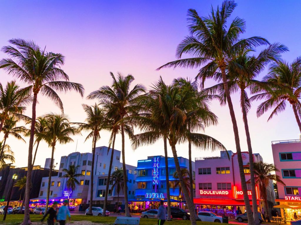 Ocean Drive a Miami Beach al crepuscolo, con alti alberi di palma in primo piano e storici hotel in stile Art Déco illuminati da insegne al neon blu e rosse.