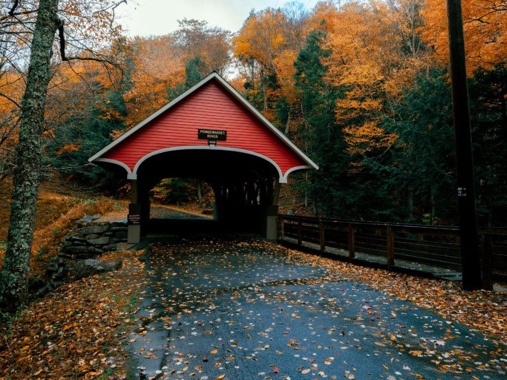 Ponte coperto di legno rosso, il Pemigewasset River Covered Bridge, circondato da alberi con foglie autunnali arancioni e una strada bagnata ricoperta di foglie secche.