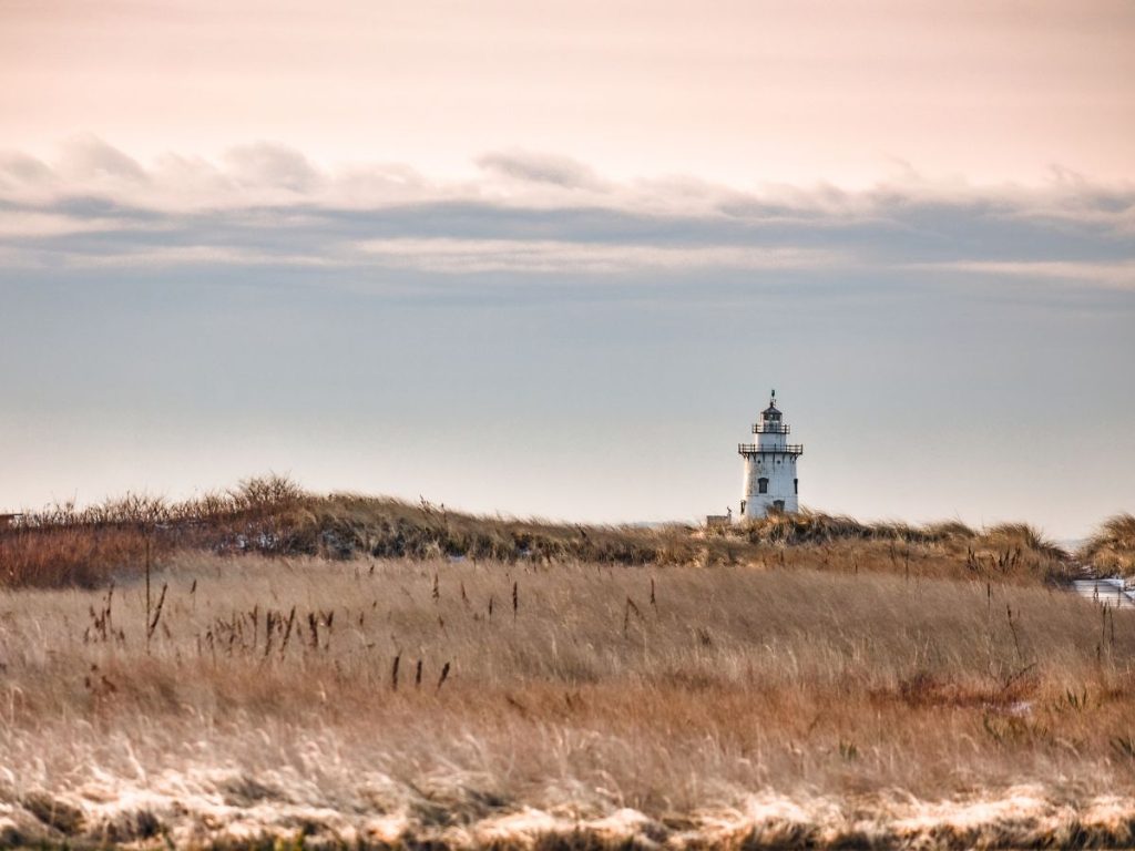 Un piccolo faro bianco e nero situato dietro dune di sabbia ed erba secca dorata, sotto un cielo di sfumature rosa e grigio al tramonto.