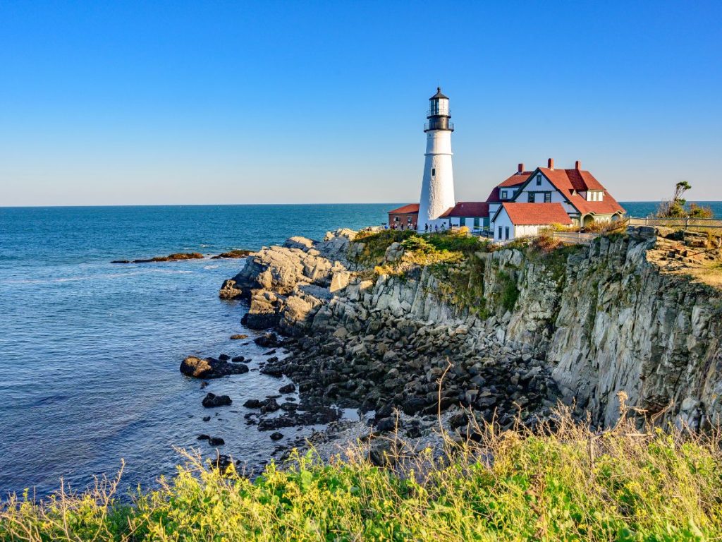 Il faro di Portland Head Light su una scogliera rocciosa a picco sul mare azzurro, affiancato da una casa d'epoca dal tetto rosso, da vedere nel New England viaggio di 12 giorni