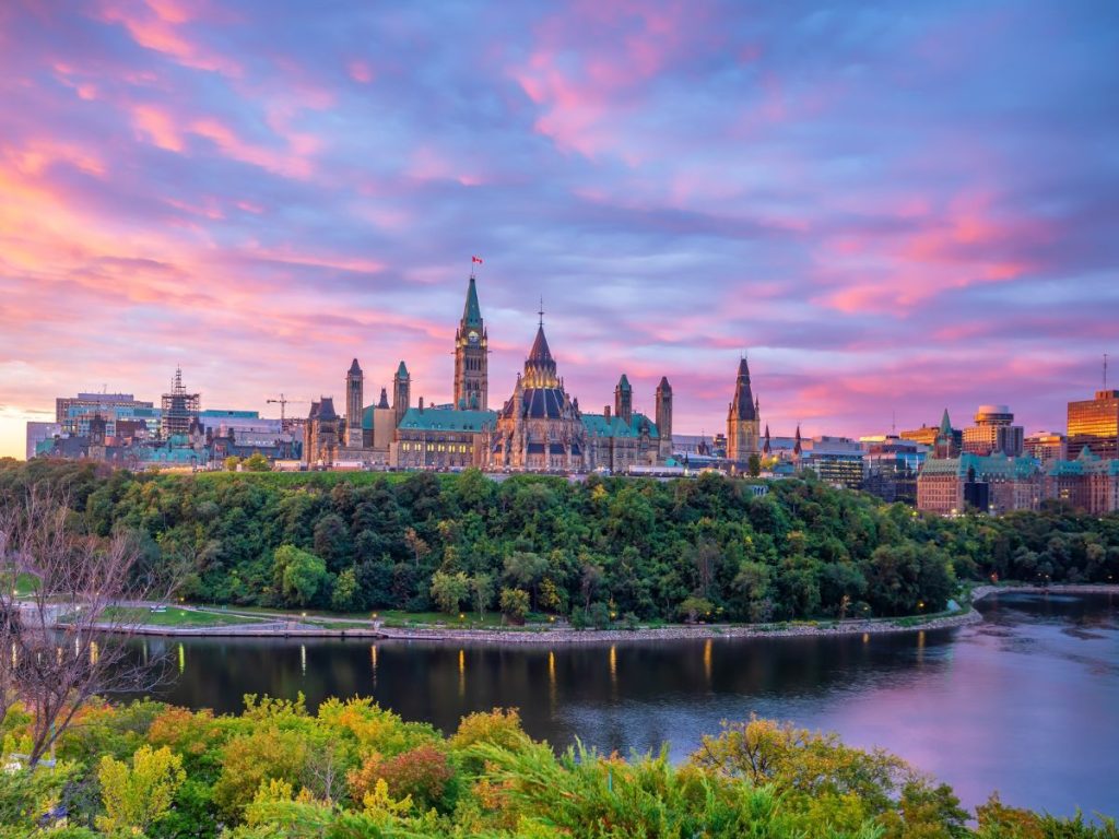 Panorama del Parlamento di Ottawa sulla collina al tramonto, circondato da vegetazione autunnale e riflesso nelle acque del fiume sotto un cielo sfumato di rosa e viola. Da vedere durante un viaggio di 15 giorni in Canada.